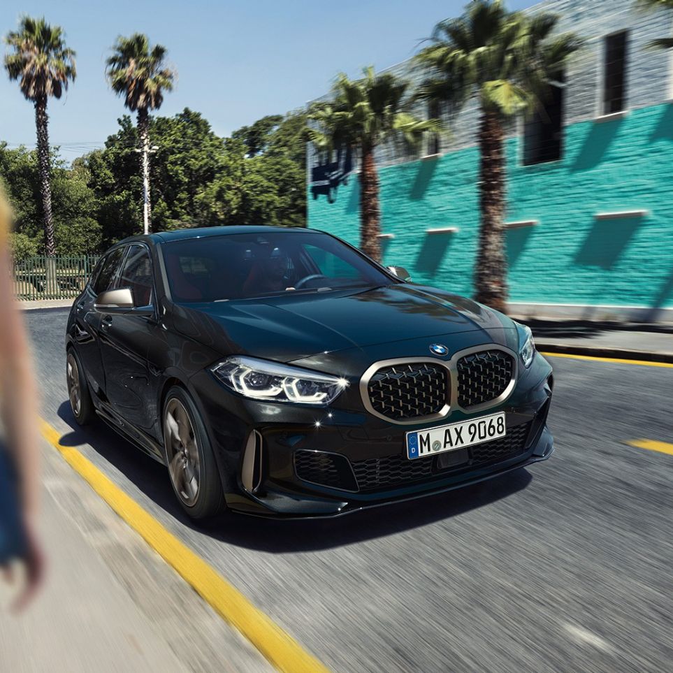 BMW M135i xDrive, vue de trois quarts avant F40. BMW M135i xDrive en Saphirschwarz metallic, vue de trois quarts avant, avec femme à l’avant-plan F40.