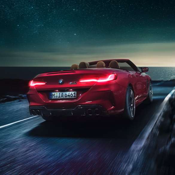 BMW M8 Competition Cabrio, vue de trois quarts arrière F91. BMW M8 Competition Cabrio en Motegi Rot metallic avec capote ouverte, vue de trois quarts arrière, roulant de nuit sur une route côtière F91.