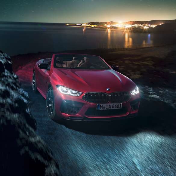 BMW M8 Competition Cabrio, vue de trois quarts face F91. BMW M8 Competition Cabrio en Motegi Rot metallic avec capote ouverte, vue de trois quarts face, roulant de nuit sur une route côtière F91.