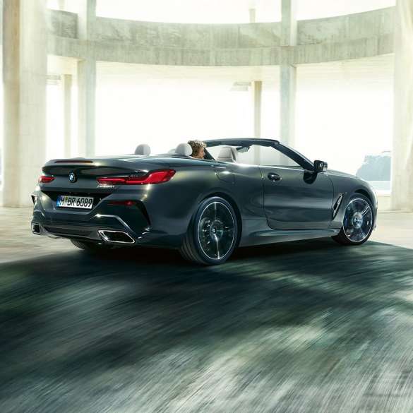 BMW M850i xDrive Cabrio, vue de trois quarts arrière G14. BMW M850i xDrive Cabrio en Dravitgrau metallic avec capote ouverte, vue de trois quarts arrière, en marche devant des immeubles en béton G14.