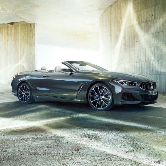 BMW M850i xDrive Cabrio, vue de trois quarts de profil G14. BMW M850i xDrive Cabrio en Dravitgrau metallic avec capote ouverte, vue de trois quarts de profil, à l’arrêt devant un immeuble en béton G14.
