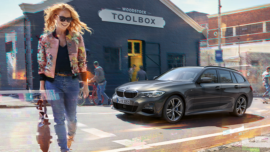 BMW 3 Reeks Touring (G21), driekwart vooraanzicht op weg met vrouw voor winkel met houten gereedschap Vrouw steekt weg over voor BMW 3 Reeks Touring (G21) en winkel met houten gereedschap.