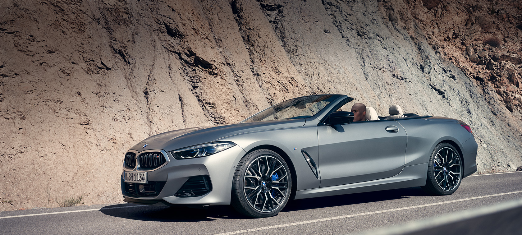 BMW 8 Reeks Cabrio G14 LCI facelift 2022 met geopende kap rijdend langs bergwand BMW 8 Reeks Cabrio G14 LCI facelift 2022 BMW Individual Frozen Pure Grey metallic zijaanzicht met geopende kap rijdend langs bergwand