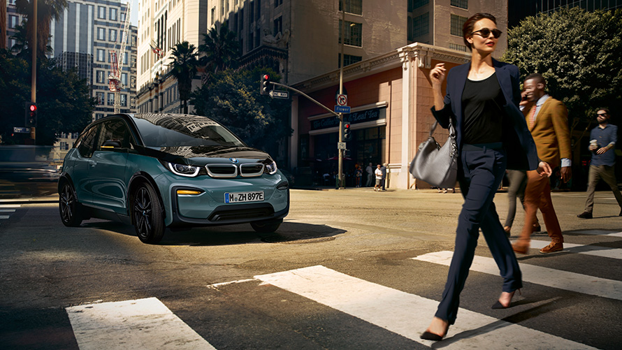 BMW i3 I01, vue de trois quarts avant avec femme qui traverse un passage piéton BMW i3 I01 Blue Ridge Mountain metallic avec accent Frozen Grey metallic, vue de trois quarts avant avec femme qui traverse un passage piéton