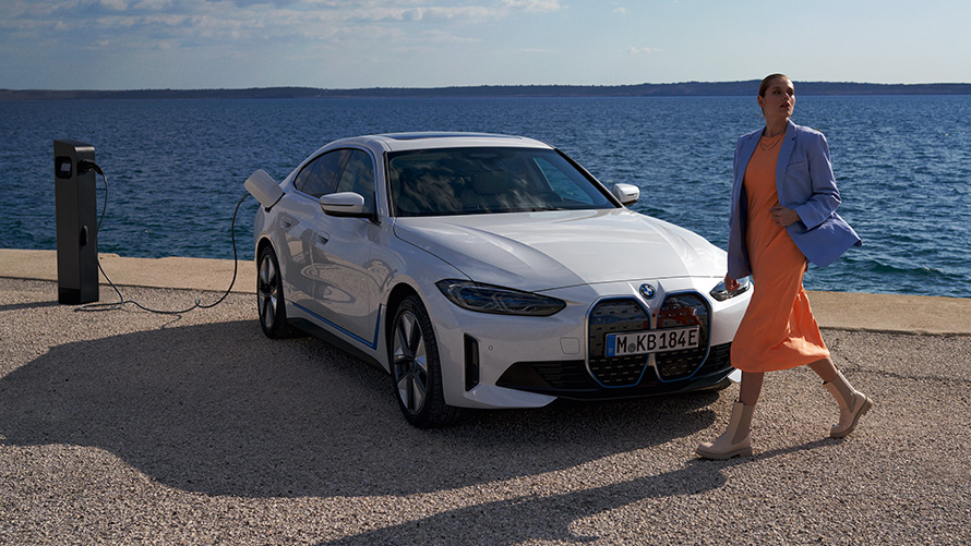 Driekwart vooraanzicht BMW i4 eDrive40 G26 2021 aan laadstation Vrouw voor BMW i4 eDrive40 G26 2021 Mineralweiß driekwart vooraanzicht aan laadpaal bij meer