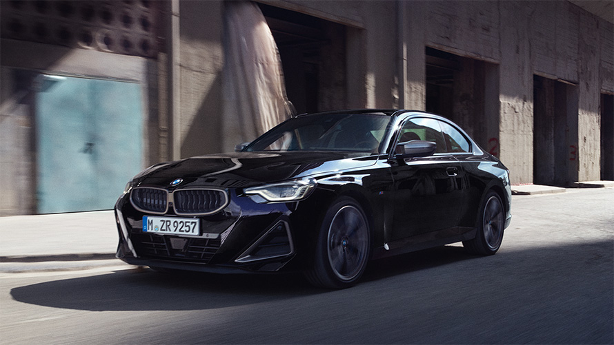 BMW M240i xDrive Coupé G42 2021 vue de trois quarts de profil en mouvement BMW M240i xDrive Coupé G42 2021 Black Sapphire metallic, vue de trois quarts de profil en mouvement devant des entrepôts