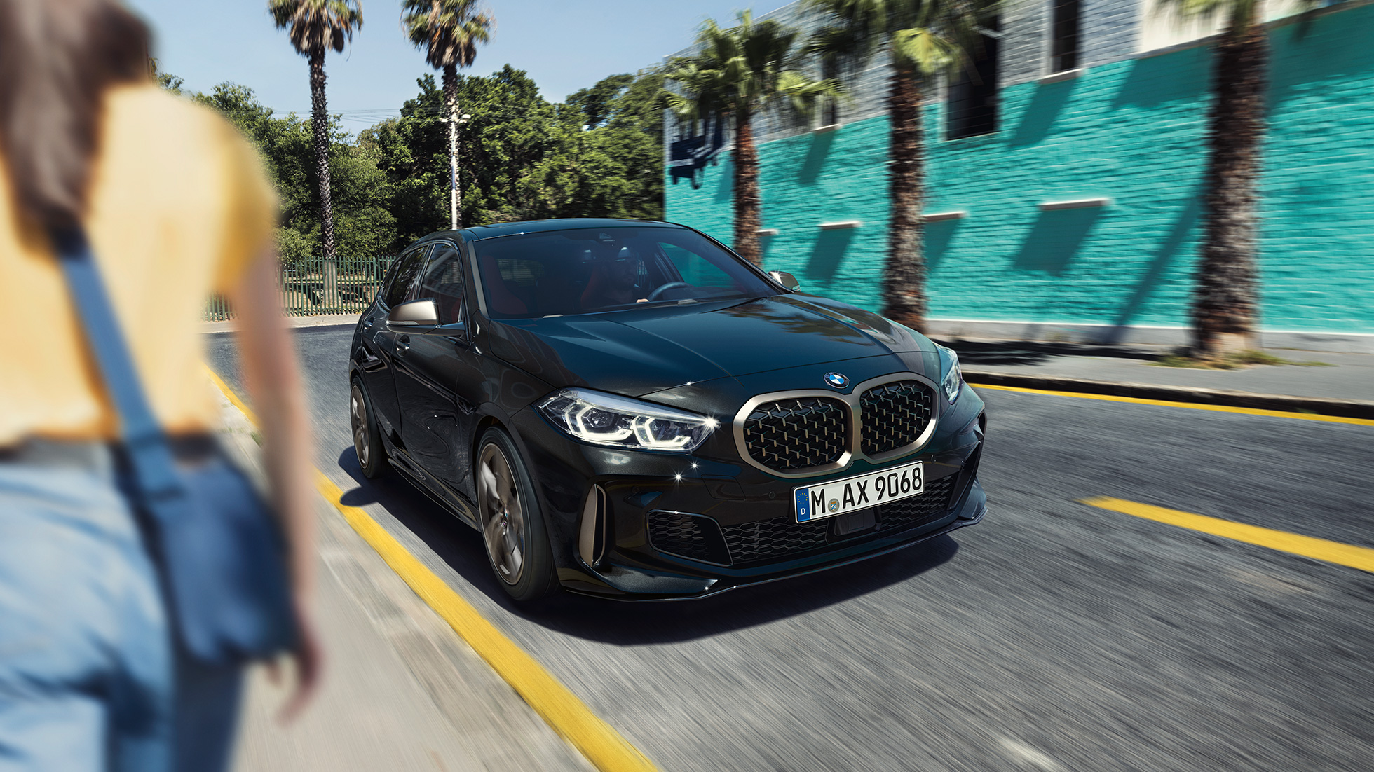 BMW M135i xDrive, vue de trois quarts avant F40. BMW M135i xDrive en Saphirschwarz metallic, vue de trois quarts avant, avec femme à l’avant-plan F40.