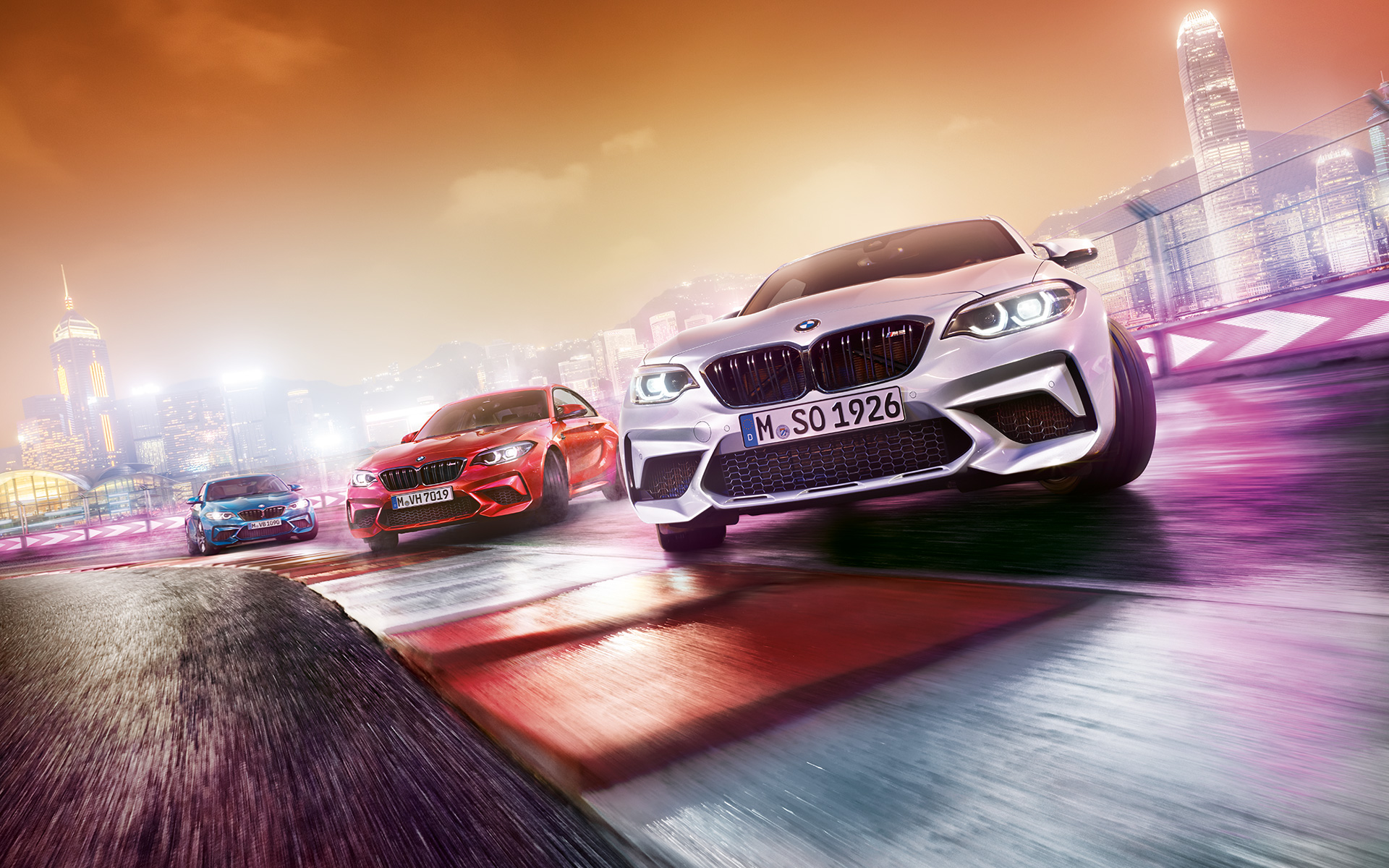 BMW M2 Competition in actie Drie BMW’s M2 Competition driften op het circuit met op de achtergrond een nachtelijke skyline.