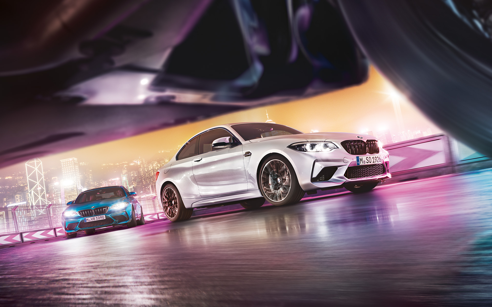 BMW M2 Competition op het weerspiegelende asfalt Twee BMW M2 Competition op het circuit vanuit kikkerperspectief met de nachtelijke grootstad als decor.