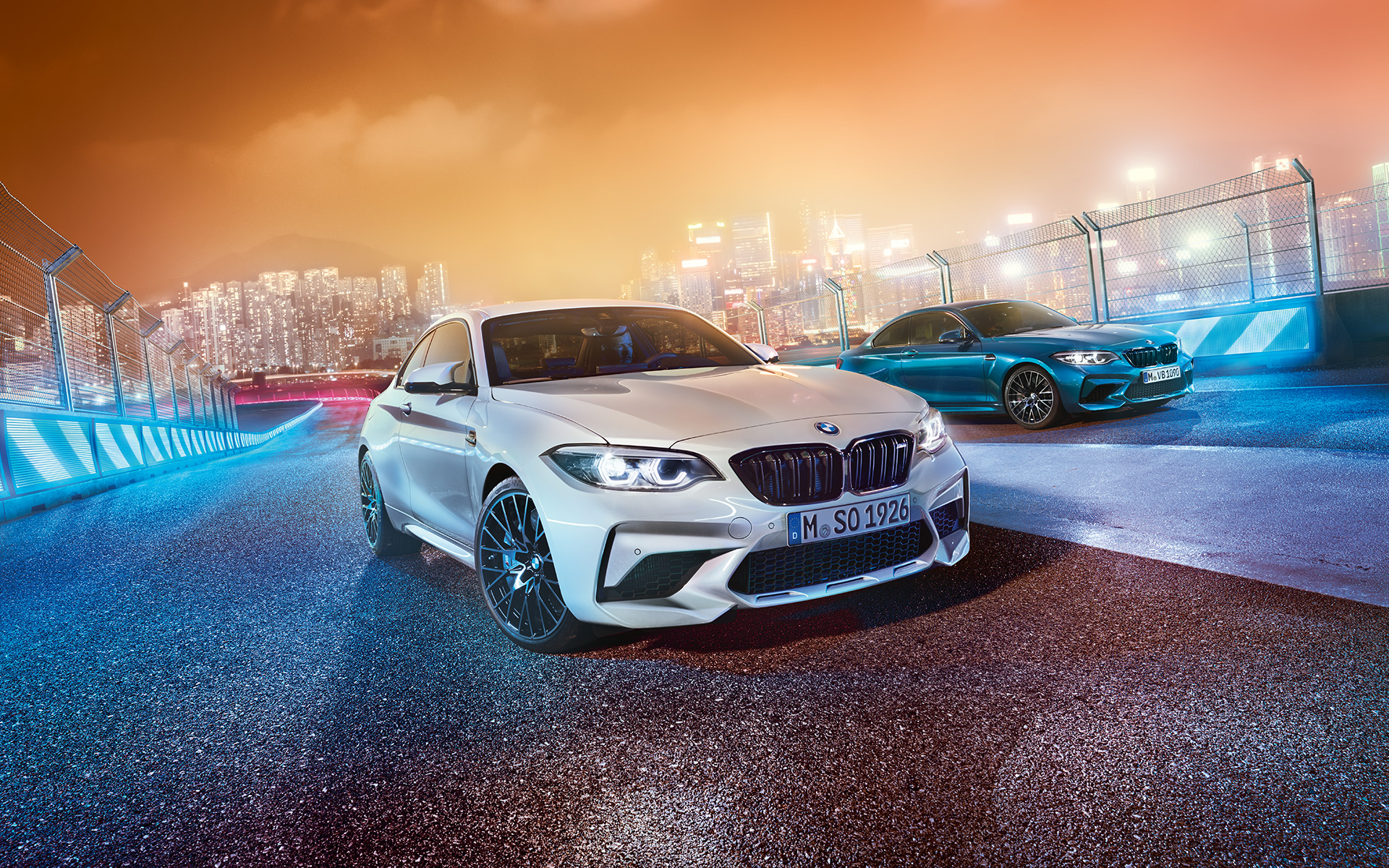 Twee BMW M2 Competition op het circuit Witte en blauwe BMW M2 Competition in schuin vooraanzicht stilstaand voor de nachtelijke grootstad op de achtergrond.