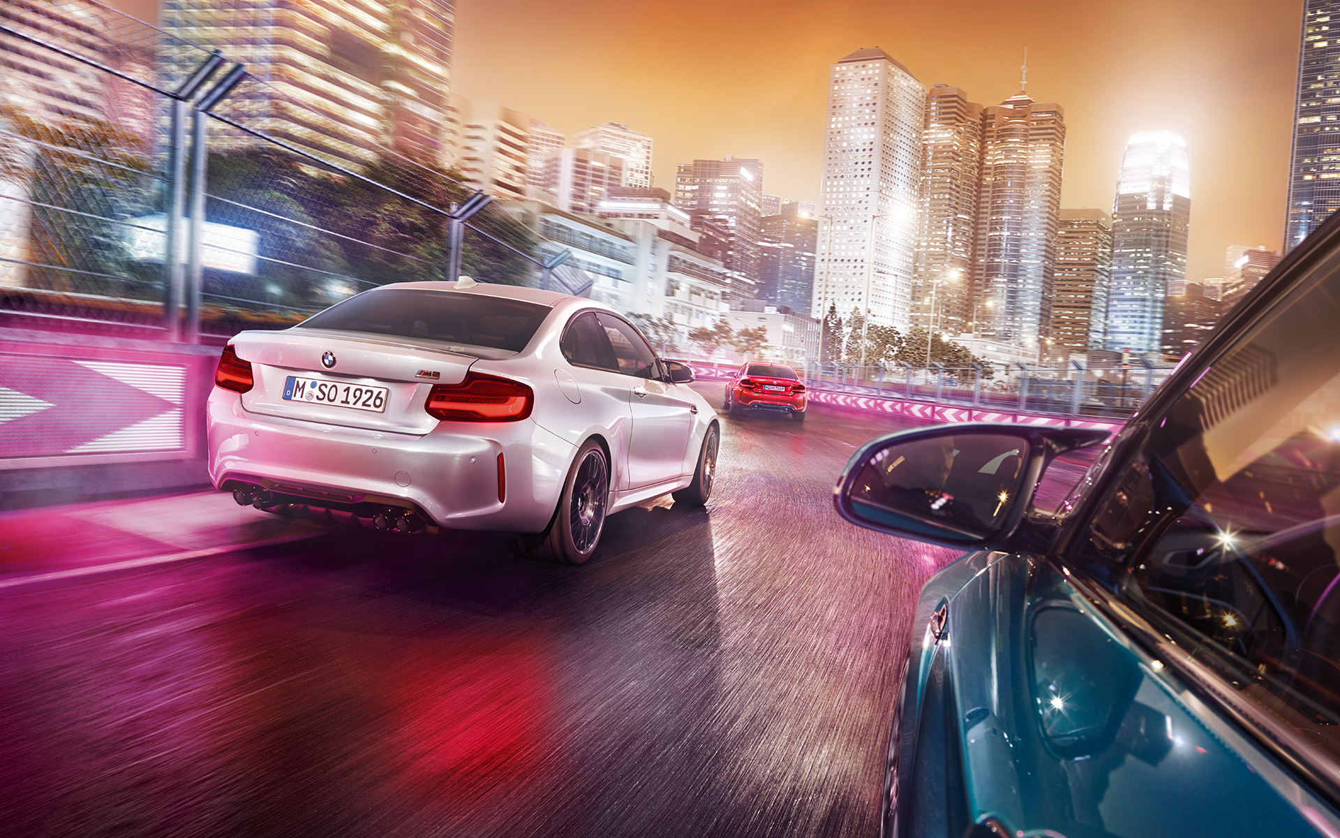 BMW M2 Competition achteraanzicht Drie BMW M2 Competition in achteraanzicht rijden op het circuit voor een oplichtende skyline.