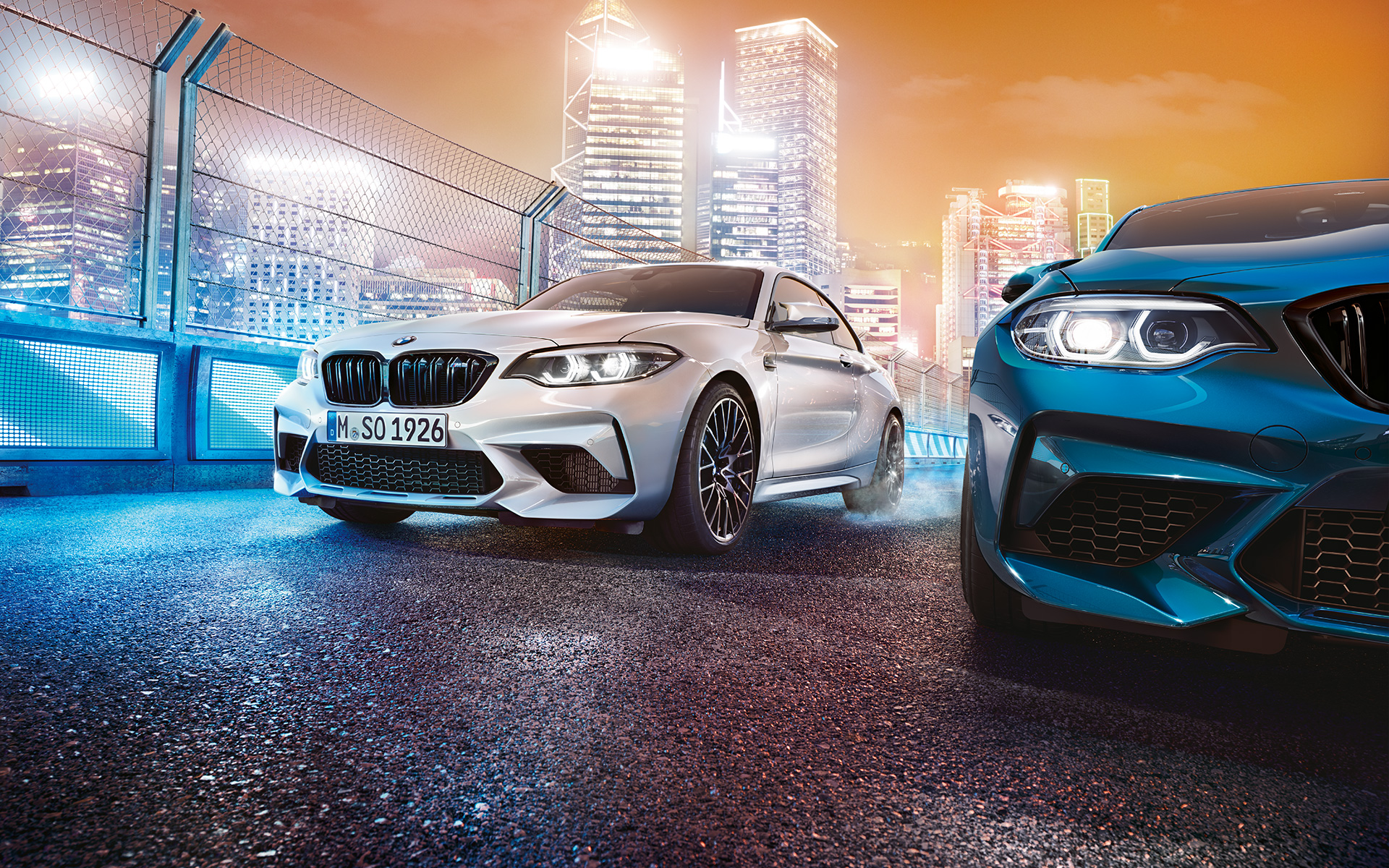 Witte en blauwe BMW M2 Competition in stilstand Witte en blauwe BMW M2 Competition in vooraanzicht staan voor de nachtelijke grootstad op de achtergrond.