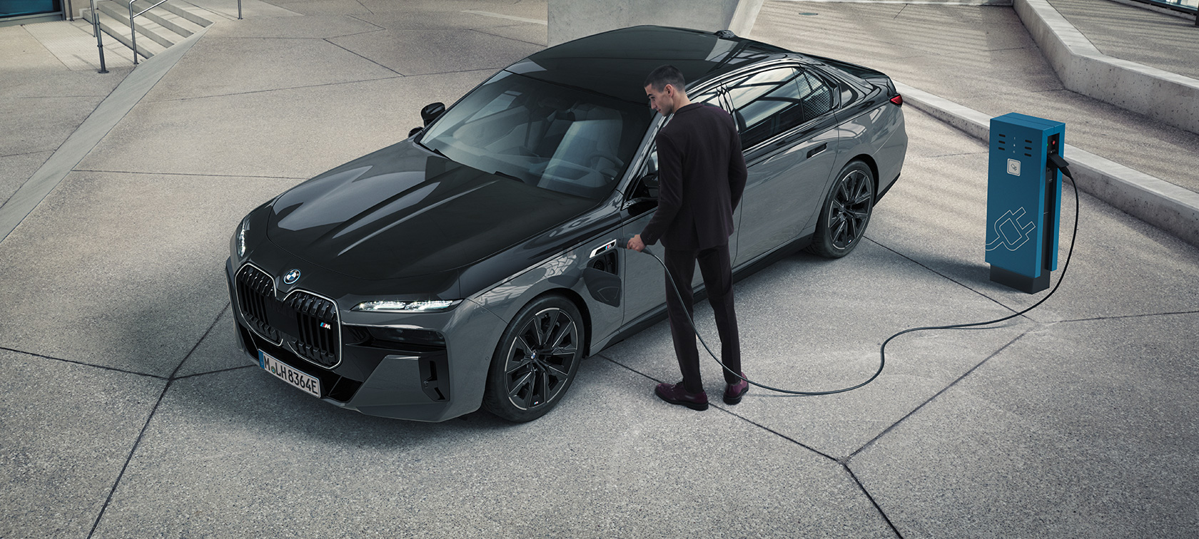BMW M760e xDrive Berline G70 pack M Performance BMW M760e xDrive Berline G70 M Performance Pack BMW teinte bi-ton BMW Individual Saphirschwarz metallic BMW Individual Dravitgrau metallic vue de trois quarts avant homme avec câble de recharge à côté d’une borne de recharge
