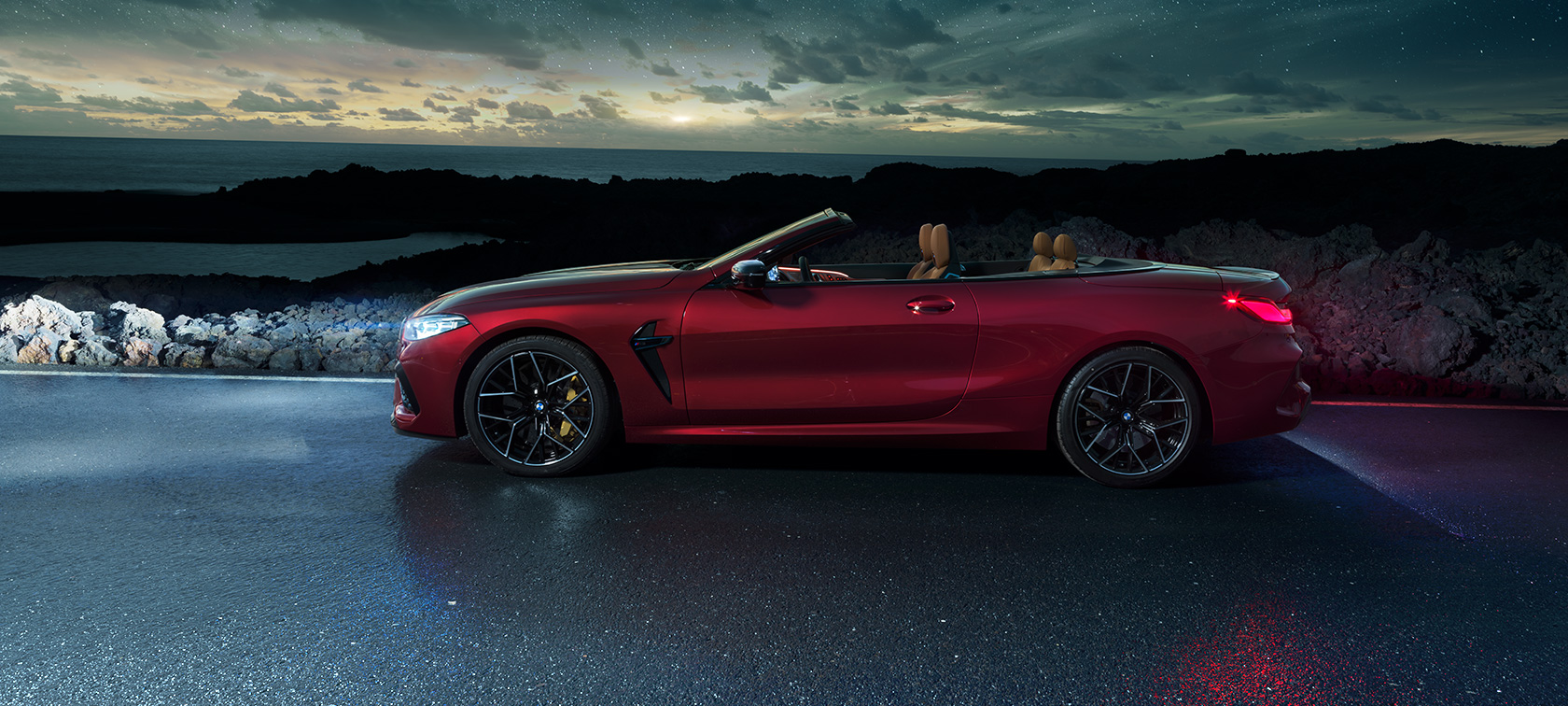 BMW M8 Competition Cabrio, vue de profil F91. BMW M8 Competition Cabrio en Motegi Rot metallic avec capote ouverte, vue de profil, à l’arrêt sur une route littorale sous un ciel étoilé F91.
