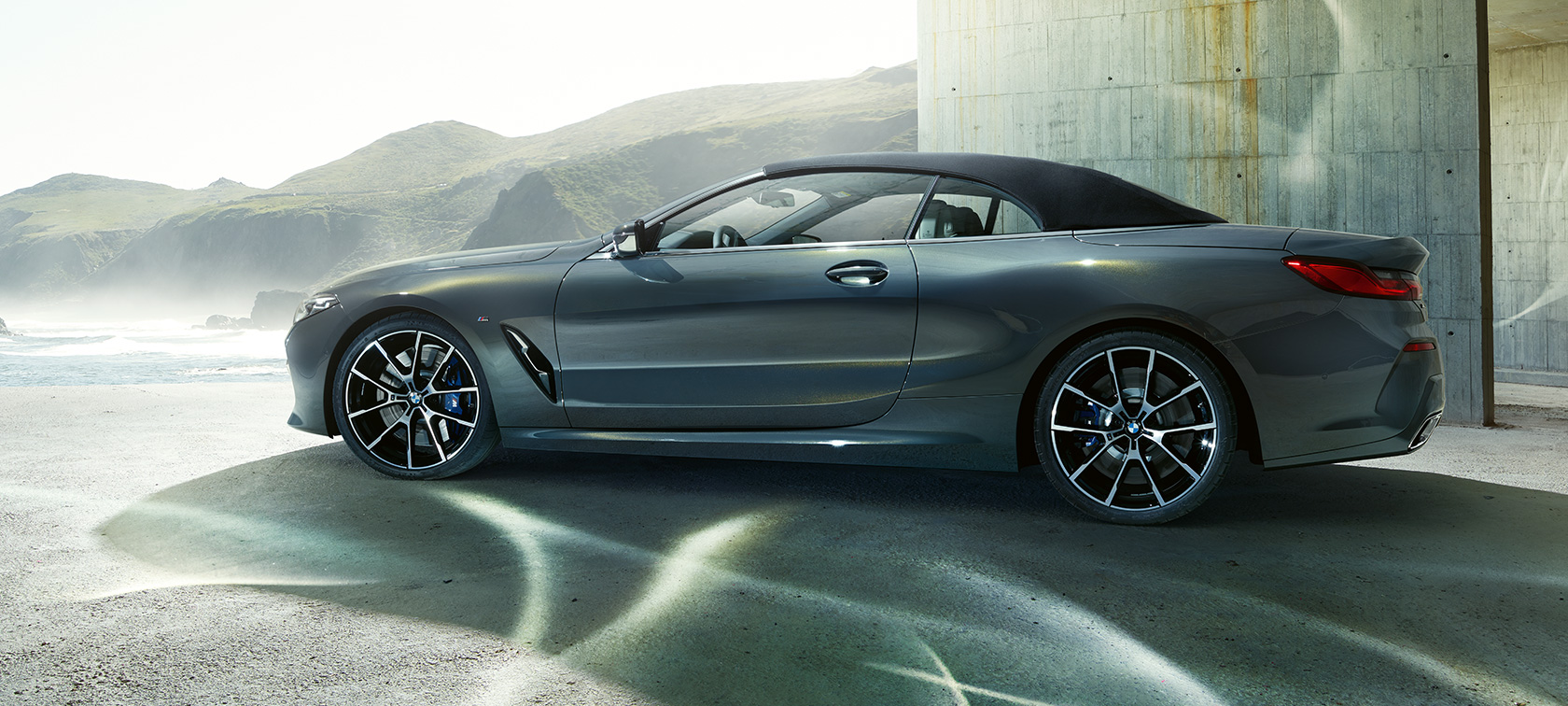 BMW M850i xDrive Cabrio, vue de profil G14. BMW M850i xDrive Cabrio en Dravit Grau metallic avec capote fermée, vue de profil devant des collines G14.