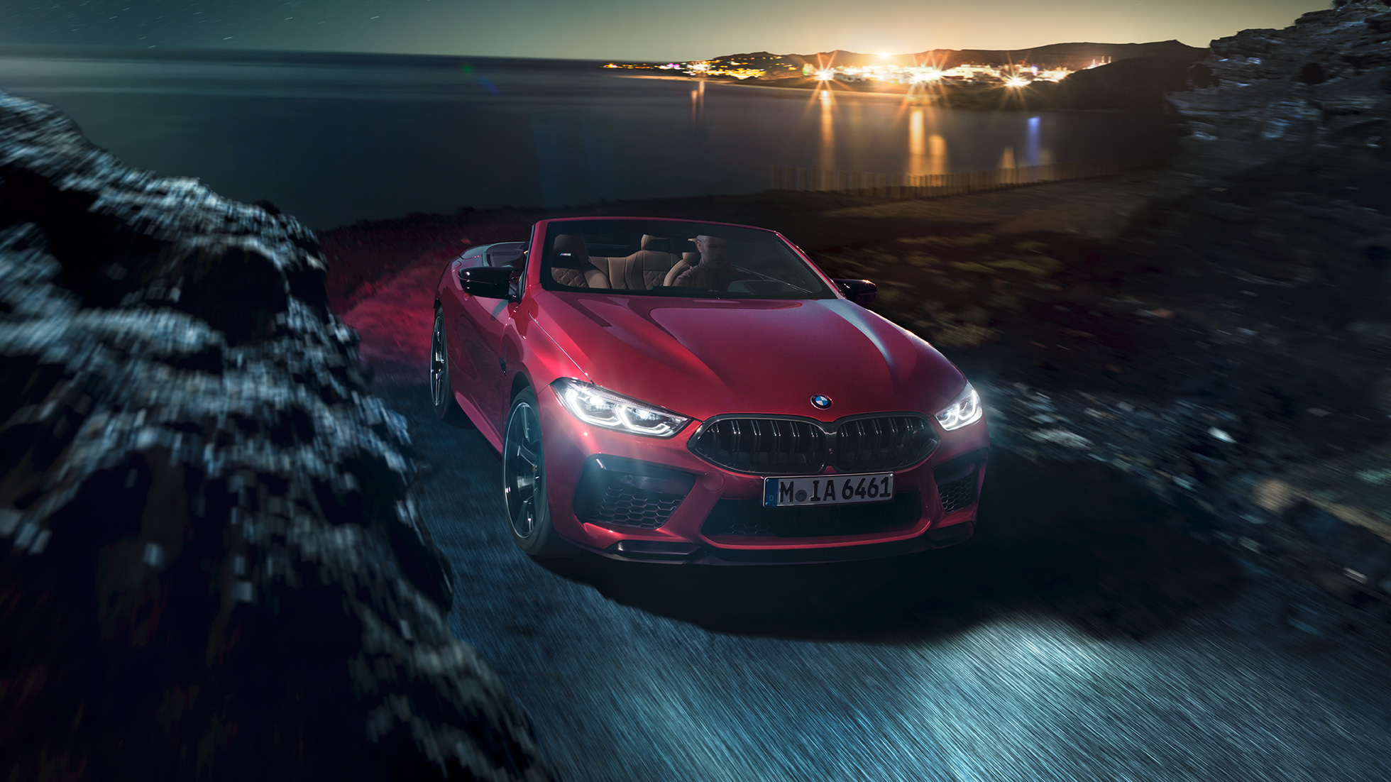 BMW M8 Competition Cabrio, vue de trois quarts face F91. BMW M8 Competition Cabrio en Motegi Rot metallic avec capote ouverte, vue de trois quarts face, roulant de nuit sur une route côtière F91.