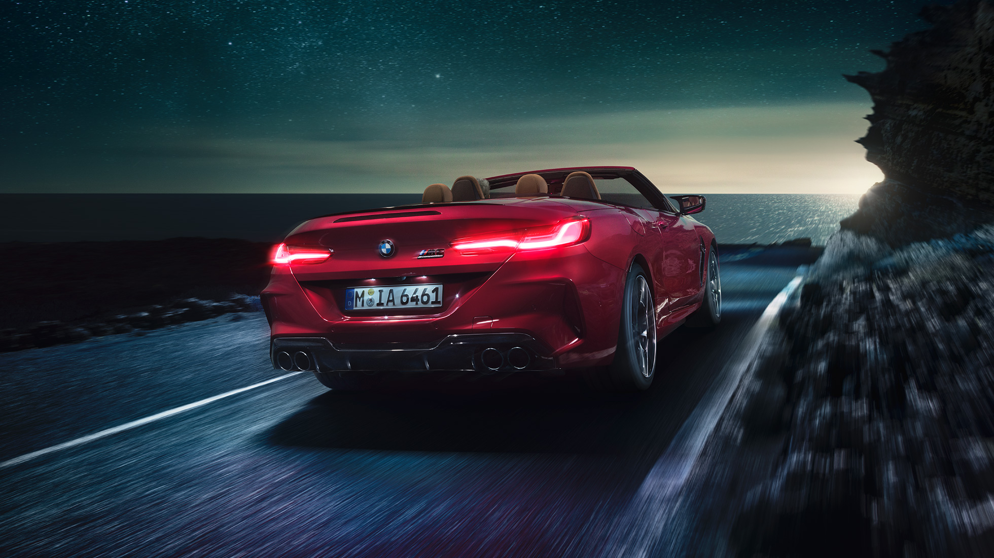 BMW M8 Competition Cabrio, vue de trois quarts arrière F91. BMW M8 Competition Cabrio en Motegi Rot metallic avec capote ouverte, vue de trois quarts arrière, roulant de nuit sur une route côtière F91.
