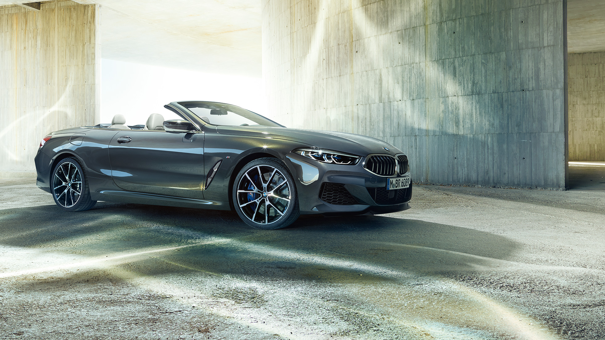 BMW M850i xDrive Cabrio, vue de trois quarts de profil G14. BMW M850i xDrive Cabrio en Dravitgrau metallic avec capote ouverte, vue de trois quarts de profil, à l’arrêt devant un immeuble en béton G14.