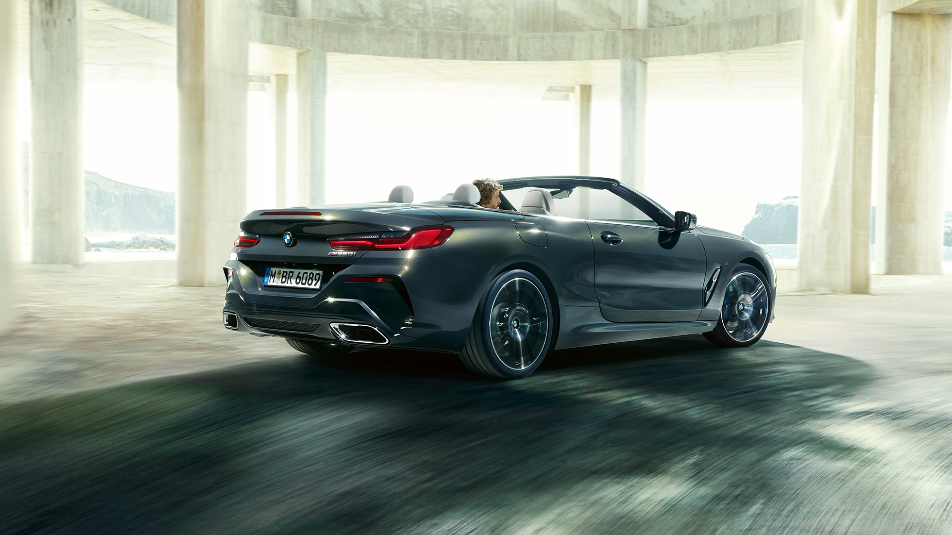 BMW M850i xDrive Cabrio, vue de trois quarts arrière G14. BMW M850i xDrive Cabrio en Dravitgrau metallic avec capote ouverte, vue de trois quarts arrière, en marche devant des immeubles en béton G14.