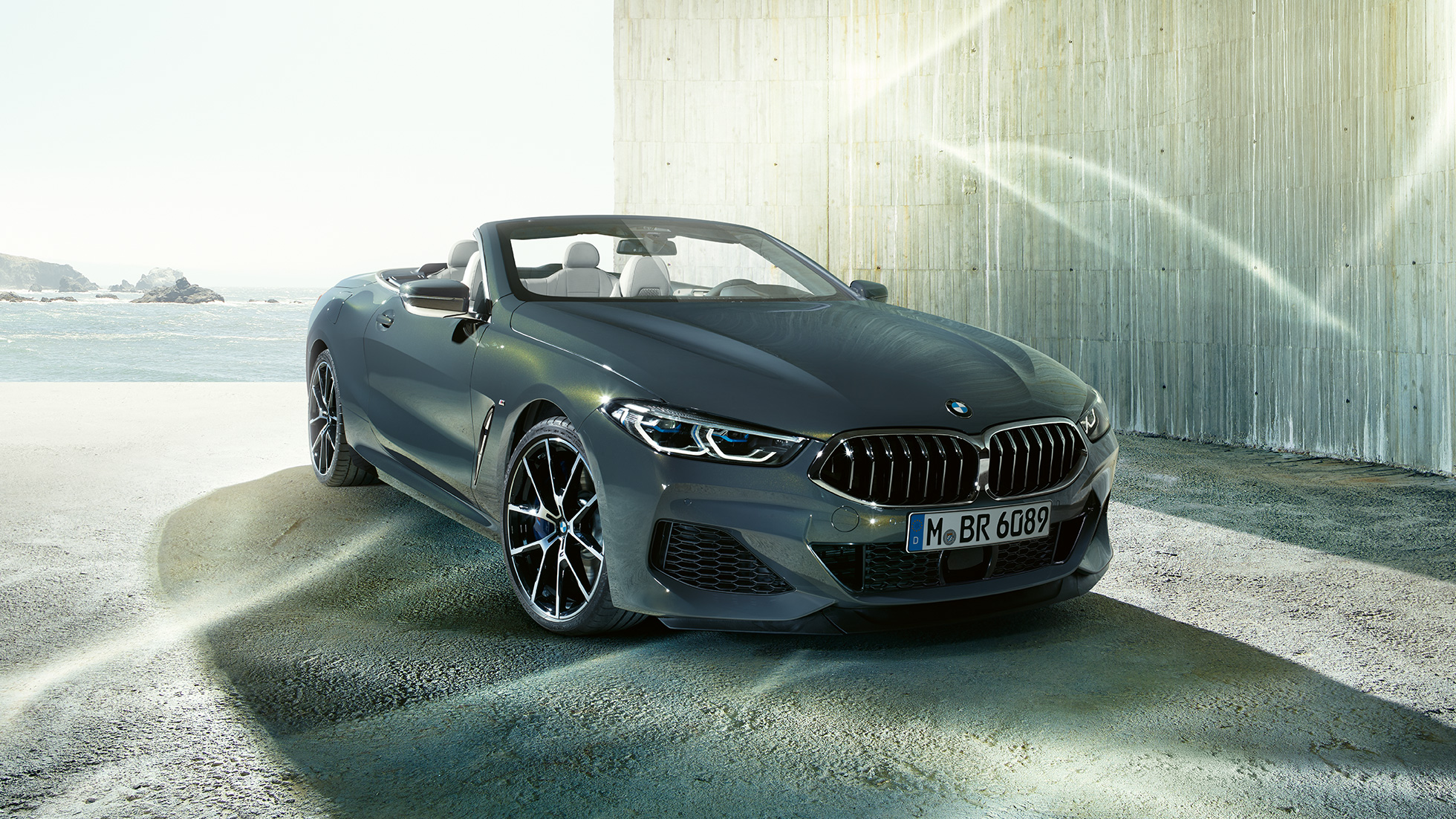 BMW M850i xDrive Cabrio, vue de trois quarts avant G14. BMW M850i xDrive Cabrio en Dravitgrau metallic avec capote ouverte, vue de trois quarts avant, à l’arrêt devant le littoral G14.