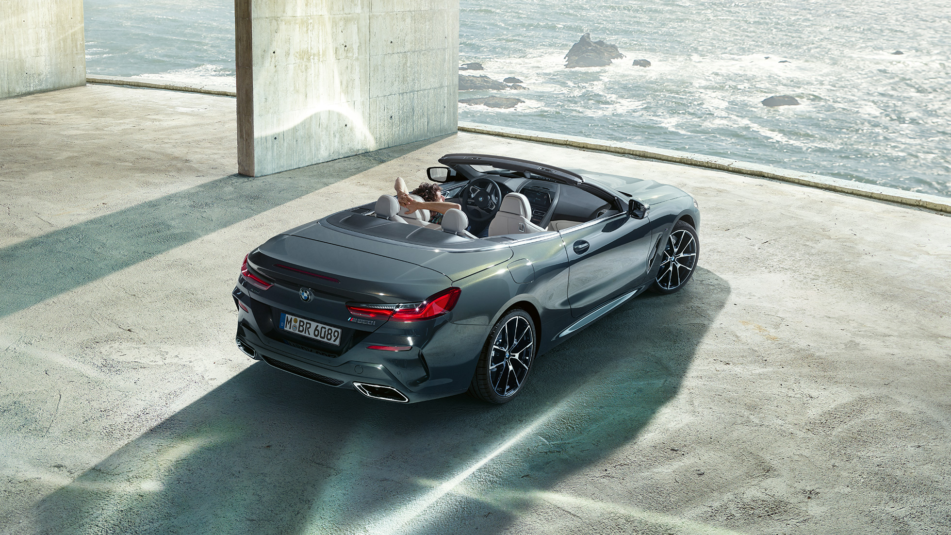 BMW M850i xDrive Cabrio, vue en plongée G14. BMW M850i xDrive Cabrio en Dravitgrau metallic avec capote ouverte, vue en plongée, en stationnement dans un bâtiment en béton devant l’océan G14.