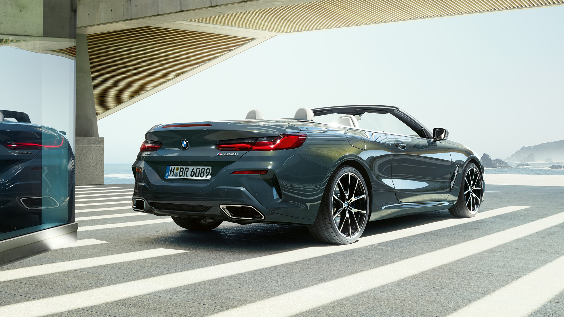 BMW M850i xDrive Cabrio, vue de trois quarts arrière G14. BMW M850i xDrive Cabrio en Dravitgrau metallic avec capote ouverte, vue de trois quarts arrière, en stationnement avec vue sur l’océan G14.