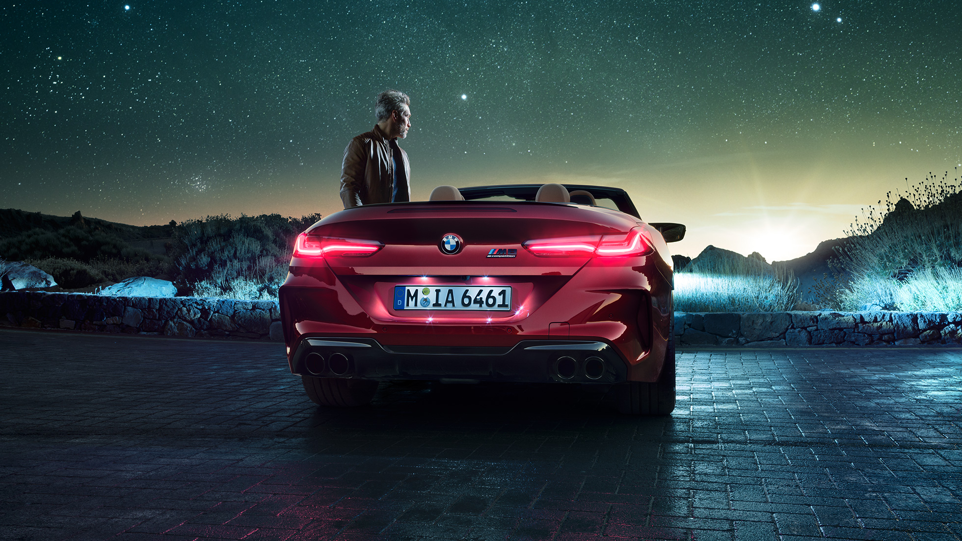 BMW M8 Competition Cabrio, vue arrière F91. BMW M8 Competition Cabrio en Motegi Rot metallic avec capote ouverte, vue arrière, à l’arrêt avec homme sous un ciel étoilé F91.