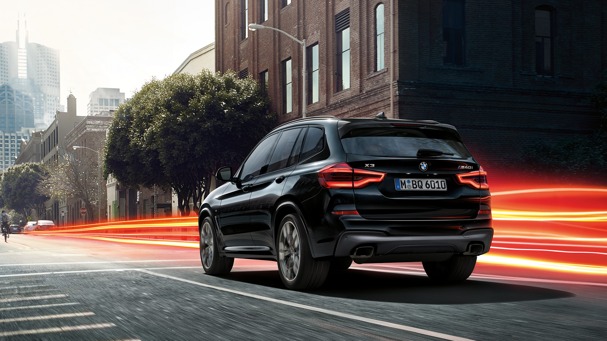 BMW X3 M40i, vue de trois quarts arrière G01. Extérieur de la BMW X3 M40i en Saphierschwarz metallic, en mouvement, vue de trois quarts arrière devant un paysage urbain avec bandes lumineuses G01