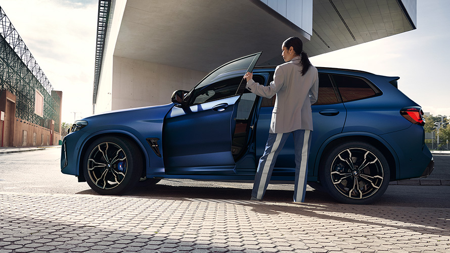 BMW X3 M Competition F97 LCI 2021 zijaanzicht met geopende deur en vrouwelijk model BMW X3 M Competition F97 LCI facelift 2021 Frozen Marina Bay Blau metallic zijaanzicht met geopende deur en vrouwelijk model