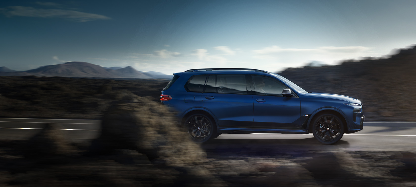 BMW X7 M60i xDrive G07 LCI Frozen Marina Bay Blue Vue de profil en mouvement sur la route devant un paysage montagneux BMW X7 M60i xDrive G07 LCI Frozen Marina Bay Blue Vue de profil en mouvement sur la route
