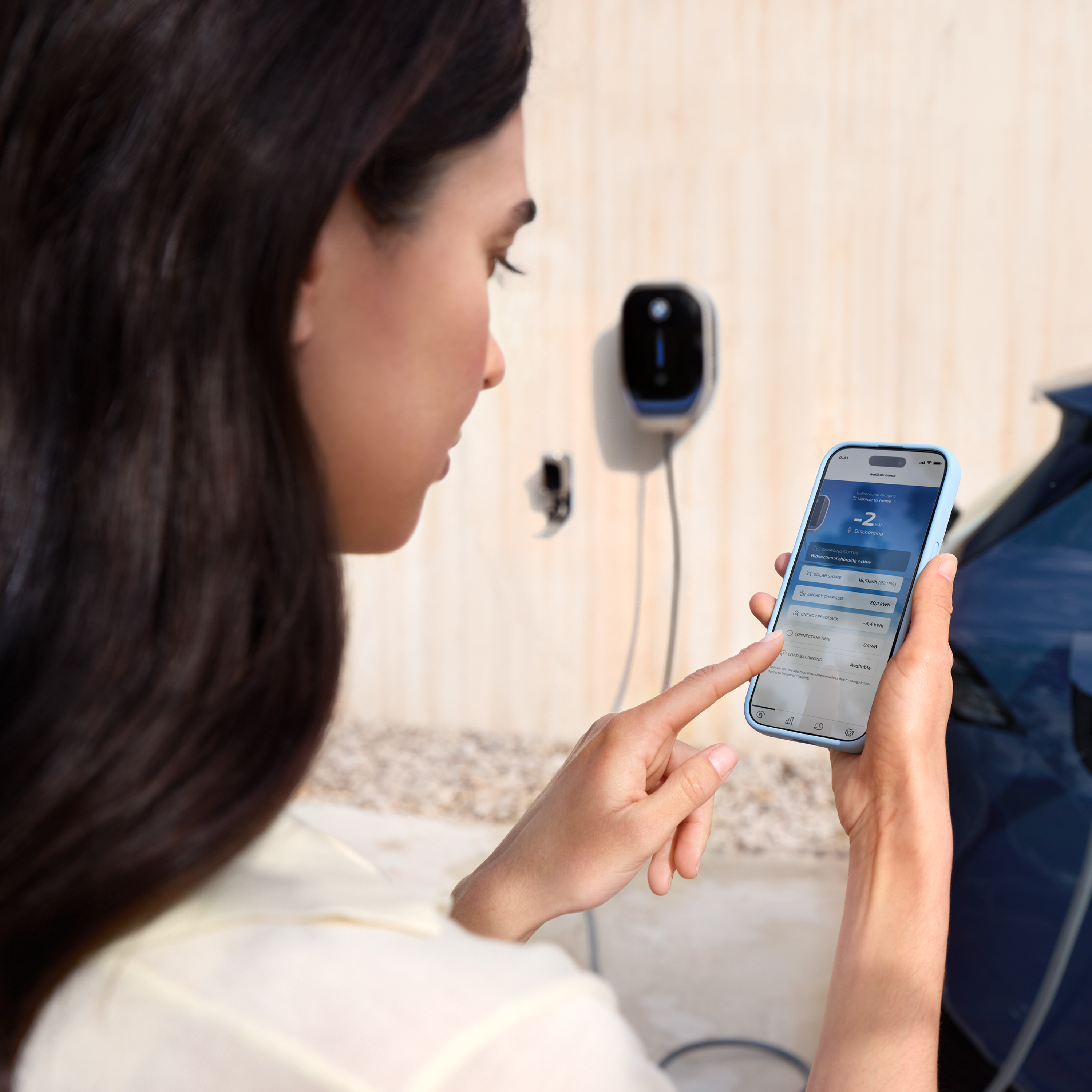 Een vrouw kijkt naar haar smartphone, op de achtergrond staat een BMW Wallbox